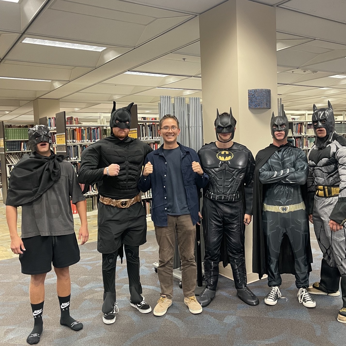 Ehsan in everyday clothes with fists up, surrounded by five people in Batman costumes in a library