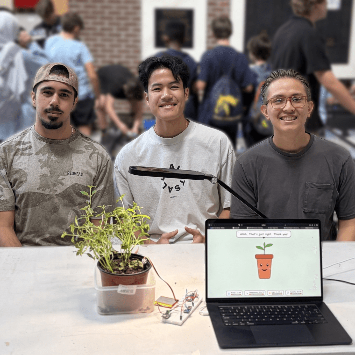 Ehsan and two teammates behind their hackathon project: a potted plant wired to sensors and a laptop showing a smiling cartoon pot
