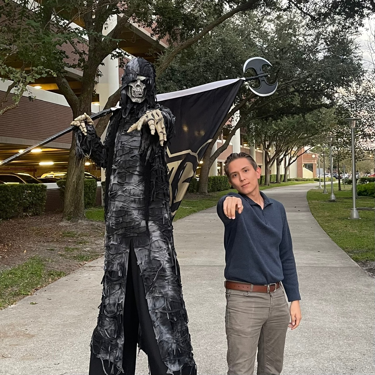 Ehsan pointing at the camera beside a towering skeleton reaper on stilts, holding a UCF flag on a battle-axe pole along a campus path